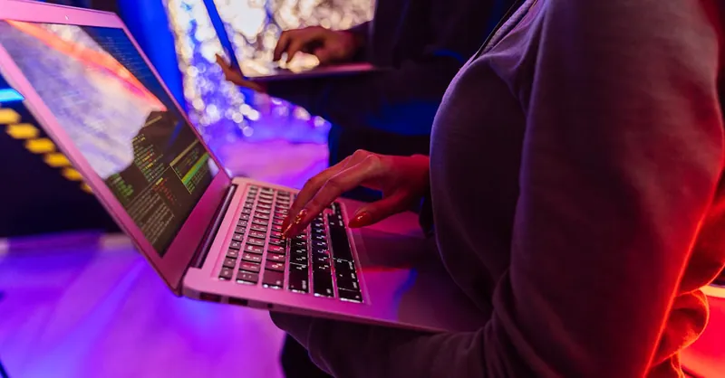 Two professionals analyzing data on laptops with colorful lighting, focusing on cybersecurity details.