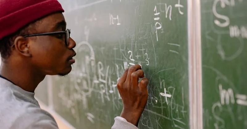 A young man with eyeglasses and a red beanie solving math problems on a chalkboard.